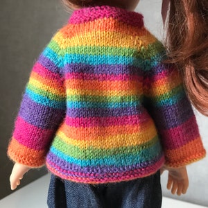 K&ouml;nnte beinhalten: Eine Puppe tr&auml;gt einen handgestrickten Regenbogenpullover. Der Pullover hat horizontale Streifen in verschiedenen Farben, darunter Rot, Orange, Gelb, Gr&uuml;n, Blau und Lila. Der Pullover hat lange &Auml;rmel und einen gerippten Kragen und B&uuml;ndchen.