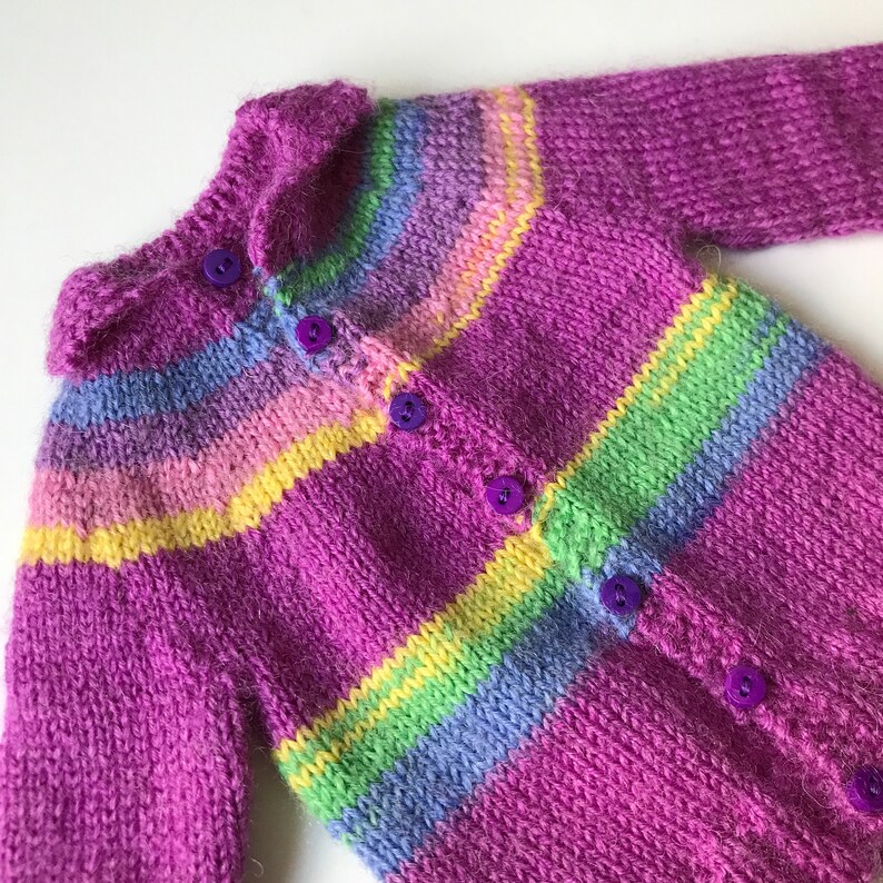 May include: A purple knitted cardigan with a colourful striped pattern. The cardigan has a collar and buttons down the front.