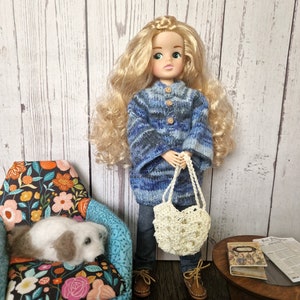 May include: A doll with long blonde hair wearing a blue and white striped sweater and jeans. She is holding a white crocheted bag. The doll is standing in front of a floral patterned armchair.