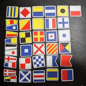 Puede incluir: Un conjunto de pegatinas de banderas de señales náuticas, cada una con un diseño único. Las banderas presentan varios colores y patrones geométricos, incluyendo rayas, cuadrados y cruces. Las pegatinas son cuadradas o rectangulares con bordes blancos.