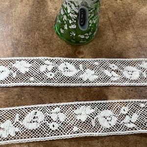 May include: A green glass thimble with a silver top sits above two strips of white lace. The lace features a floral pattern. The thimble has a decorative design with a black and white swirl. The background is a brown wooden surface.
