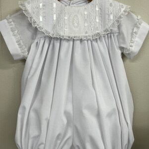 May include: White baby bubble romper with a lace collar and short sleeves. The collar has a satin ribbon trim and a delicate floral embroidery.