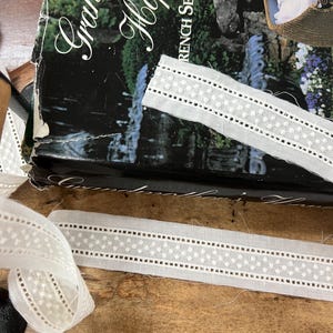 May include: Close-up of white lace ribbon with a dotted and eyelet pattern, lying on a wooden surface. The ribbon is next to a book titled "Grandma's Hope Chest" with the text "French Sewing by..." visible.