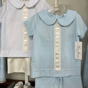 May include: Two baby boy outfits, one white and one light blue, with Peter Pan collars and short sleeves. Both outfits have a white ribbon detail down the front. The outfits are hanging on hangers and there are two pairs of white and brown shoes on a white stand.