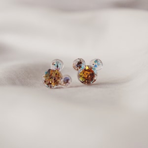 May include: A pair of clear resin stud earrings with gold and blue glitter embedded inside. The earrings are shaped like Mickey Mouse ears.