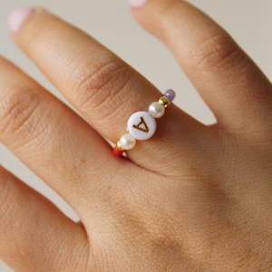 Pode incluir: Um anel de contas com um charme de letra 'A' branca, incluindo uma mistura de contas roxas, brancas e douradas.