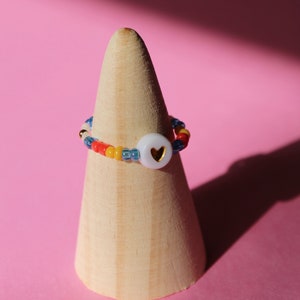 Pode incluir: Um anel de contas coloridas com um charme de coração branco. O anel é exibido em um suporte de anel de madeira.