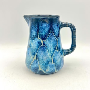 Op de afbeelding: Blauwe keramische kan met een bamboe-stijl handvat. De kan is geglazuurd in blauwtinten, met een patroon van overlappende vormen. De rand en het handvat zijn donkerblauw. Het heeft een schenktuit.