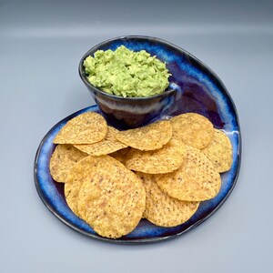 May include: A dark blue ceramic serving dish with a built-in bowl filled with guacamole. The dish is filled with tortilla chips. The dish has a unique, swirled design. The guacamole is bright green.