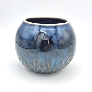 Peut inclure: Un vase rond en céramique bleue avec une finition brillante. Le vase a un intérieur bleu foncé et un extérieur bleu clair texturé avec des accents dorés près de la base. Le vase est sur fond blanc.
