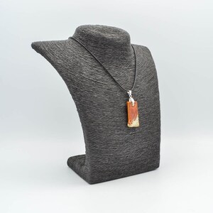 Op de afbeelding: Een rechthoekige hanger van bruin en gouden hars met zilveren accenten hangt aan een zwart koord op een zwarte sieradenstandaard.