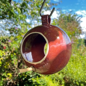 Può includere: Mangiatoia per uccelli sferica in ceramica, con una profonda glassa marrone-rossastra. Presenta una grande apertura circolare e un piccolo posatoio in legno. Appesa a un gancio di metallo nero, è ideale per attirare gli uccelli nel tuo giardino.