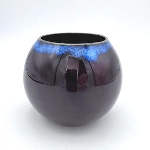 Op de afbeelding: Een ronde, donkerpaarse keramische vaas met een glanzende afwerking. De bovenrand heeft een blauwe glazuurlaag. De vaas reflecteert licht, waardoor highlights op het oppervlak ontstaan. De binnenkant is zwart.
