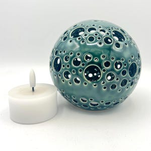 Op de afbeelding: Een bolvormige, blauwgroene keramische kaarsenhouder met een getextureerd oppervlak met talloze ronde uitsparingen van verschillende groottes, vergezeld van een klein wit cilindrisch led-theelichtje met een puntige vlamvorm.