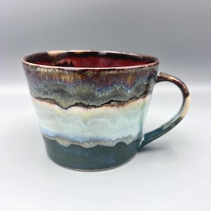 May include: A ceramic mug featuring a wide, flared rim and a single handle. The exterior glaze displays horizontal bands of colour, transitioning from a deep dark blue at the base to light blue, cream, and a dark, mottled brown at the top rim.