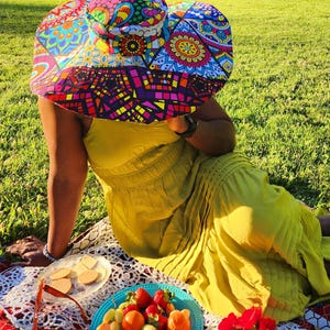 May include: A person wearing a large, colorful, patterned sun hat sits on a blanket in a grassy park. The hat has a wide brim and is made of fabric with a patchwork design. The person is wearing a yellow dress and has a plate of fruit and snacks in front of them.