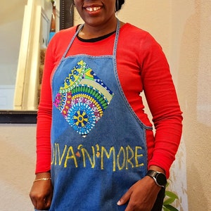 May include: A woman wearing a red long-sleeved shirt and a denim apron with a colorful geometric design and the text "Diva N' More". She is also wearing a black and green patterned head wrap.