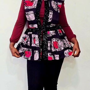 May include: A sleeveless, patterned top with a high collar and a flared waist. The top features a black background with pink and red abstract shapes. A black sequined strip runs down the front. The top is worn over a long-sleeved burgundy shirt.