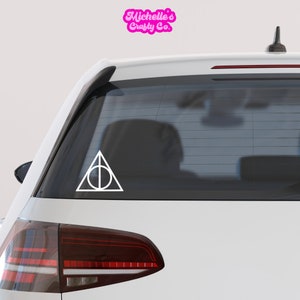 May include: White vinyl decal of the Deathly Hallows symbol from the Harry Potter series on the back window of a white car.