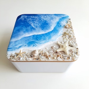 Può includere: Una scatola quadrata bianca con un design a onda oceanica in resina blu e bianca sulla parte superiore. La scatola è decorata con conchiglie e stelle marine.