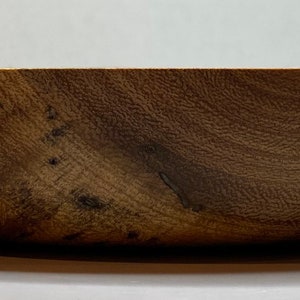 Puede incluir: Un cuenco de madera con un acabado natural de vetas. El cuenco tiene un dise&ntilde;o ancho y poco profundo y un interior liso y curvado. La madera tiene un color marr&oacute;n c&aacute;lido con vetas y nudos m&aacute;s oscuros, lo que le da un aspecto r&uacute;stico. El cuenco probablemente se utiliza para servir comida o como pieza decorativa.