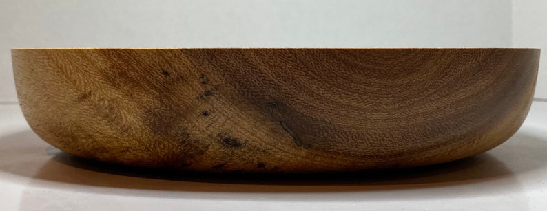 Puede incluir: Un cuenco de madera con un acabado natural de vetas. El cuenco tiene un dise&ntilde;o ancho y poco profundo y un interior liso y curvado. La madera tiene un color marr&oacute;n c&aacute;lido con vetas y nudos m&aacute;s oscuros, lo que le da un aspecto r&uacute;stico. El cuenco probablemente se utiliza para servir comida o como pieza decorativa.