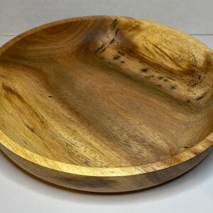 Puede incluir: Un cuenco redondo de madera con un acabado de vetas naturales. El cuenco tiene poca profundidad y un borde ancho. La madera presenta diferentes tonos de marr&oacute;n claro y oscuro. El cuenco probablemente se utiliza para servir alimentos o como pieza decorativa.