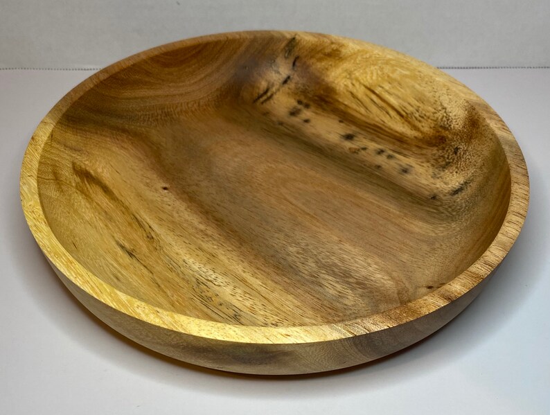 Puede incluir: Un cuenco redondo de madera con un acabado de vetas naturales. El cuenco tiene poca profundidad y un borde ancho. La madera presenta diferentes tonos de marr&oacute;n claro y oscuro. El cuenco probablemente se utiliza para servir alimentos o como pieza decorativa.