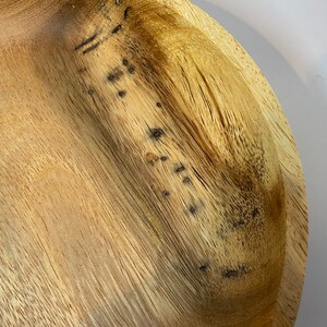 Puede incluir: Primer plano de un cuenco de madera de color marr&oacute;n claro con vetas visibles. El cuenco tiene una forma poco profunda y redondeada, con algunas manchas oscuras. La veta de la madera es prominente, creando una est&eacute;tica natural y r&uacute;stica.