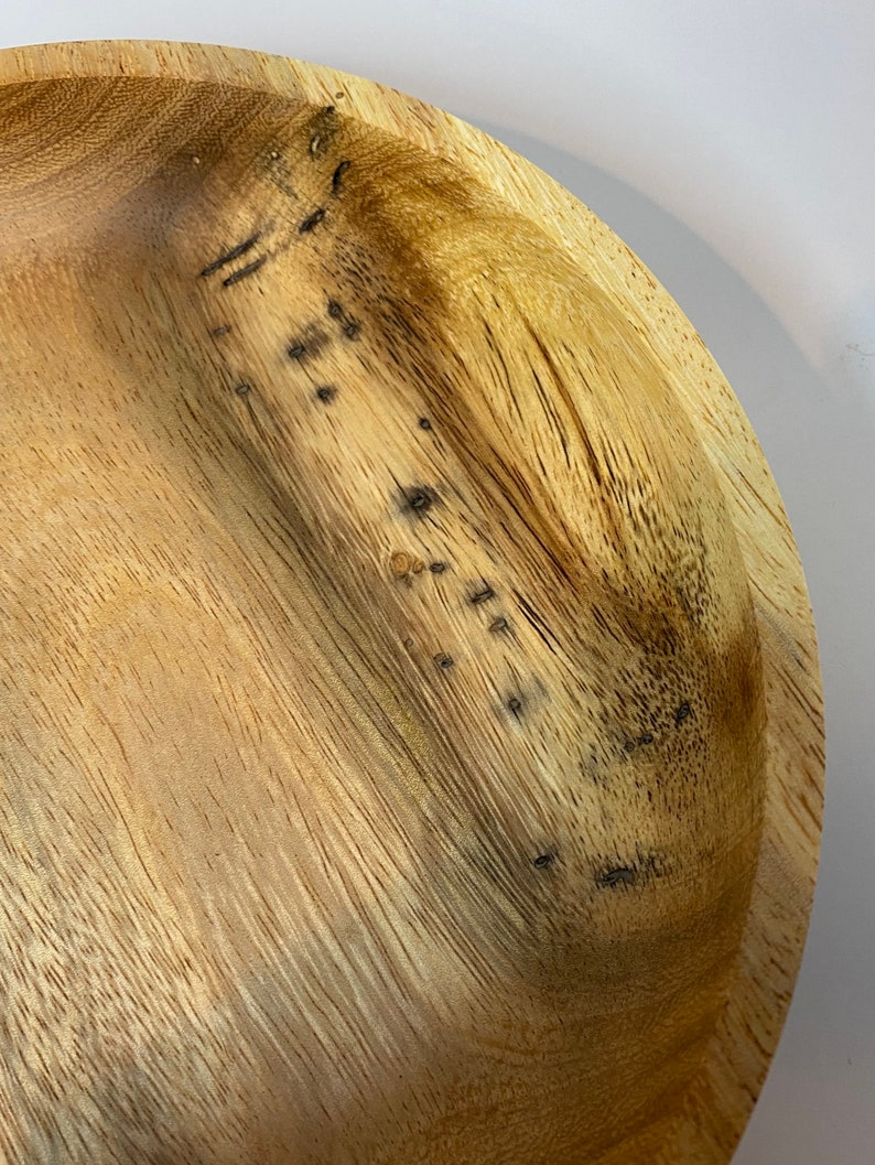 Puede incluir: Primer plano de un cuenco de madera de color marr&oacute;n claro con vetas visibles. El cuenco tiene una forma poco profunda y redondeada, con algunas manchas oscuras. La veta de la madera es prominente, creando una est&eacute;tica natural y r&uacute;stica.