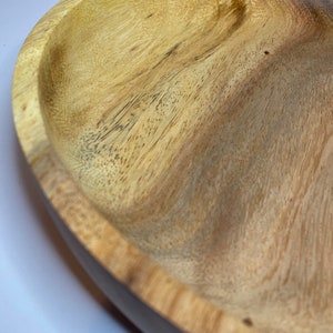 Puede incluir: Un cuenco de madera con un patr&oacute;n de vetas naturales. El cuenco tiene una forma poco profunda y redondeada con un borde oscuro. La madera es de color marr&oacute;n claro con vetas y nudos m&aacute;s oscuros, mostrando la textura natural.