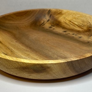 Puede incluir: Un cuenco redondo de madera con un acabado natural de vetas. El cuenco tiene poca profundidad y un borde ancho. La madera tiene diferentes tonos de marr&oacute;n y beige, con patrones de vetas visibles. Este cuenco es adecuado para servir alimentos o como objeto decorativo.