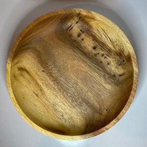 Puede incluir: Un cuenco redondo de madera con un patr&oacute;n de vetas naturales. El cuenco tiene poca profundidad y una superficie lisa y pulida. La madera presenta variaciones de color, que van del beige claro al marr&oacute;n m&aacute;s oscuro.