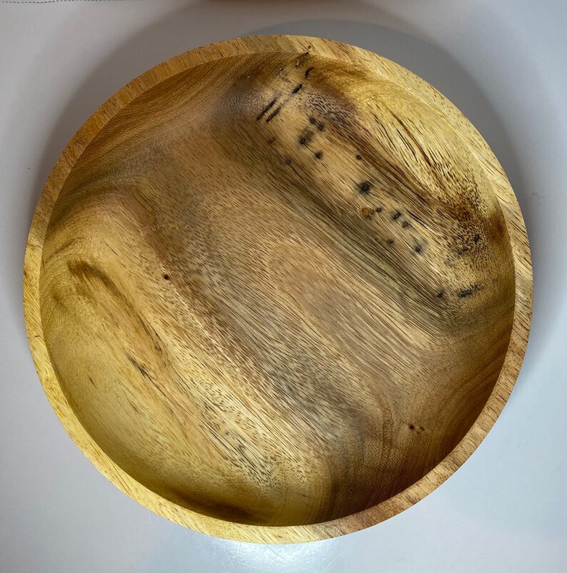 Puede incluir: Un cuenco redondo de madera con un patr&oacute;n de vetas naturales. El cuenco tiene poca profundidad y una superficie lisa y pulida. La madera presenta variaciones de color, que van del beige claro al marr&oacute;n m&aacute;s oscuro.