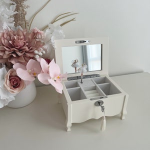 May include: Cream-coloured jewellery box with a mirrored lid, open to reveal compartments. A ballerina figurine is reflected in the mirror. The box has a key and tassel. Decorative flowers in a white vase are in the background.