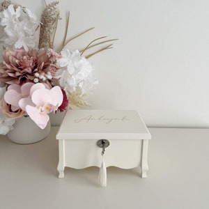 May include: A cream-coloured wooden jewellery box with the name "Anliyah" in gold script on the lid. The box has a key lock with a tassel and decorative legs. A vase of flowers is in the background.