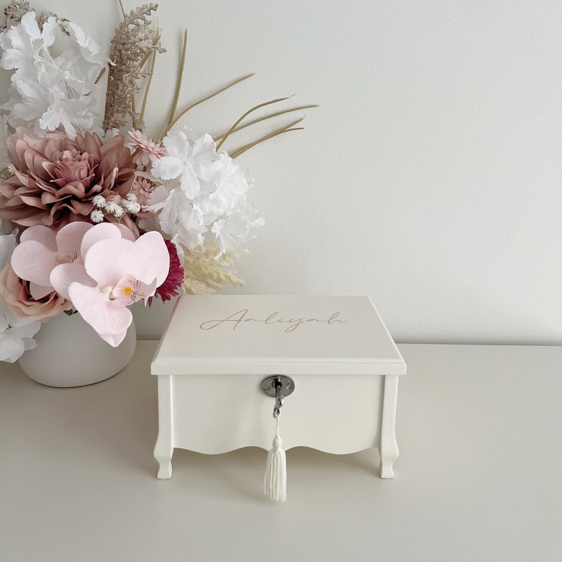 May include: A cream-coloured wooden jewellery box with the name "Anliyah" in gold script on the lid. The box has a key lock with a tassel and decorative legs. A vase of flowers is in the background.