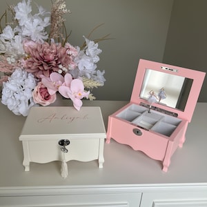 May include: Two jewellery boxes, one cream and one pink, are on a white surface. The cream box has the name "Anliyah" in cursive. The pink box has a mirror and a ballerina figurine. A bouquet of flowers is in the background.