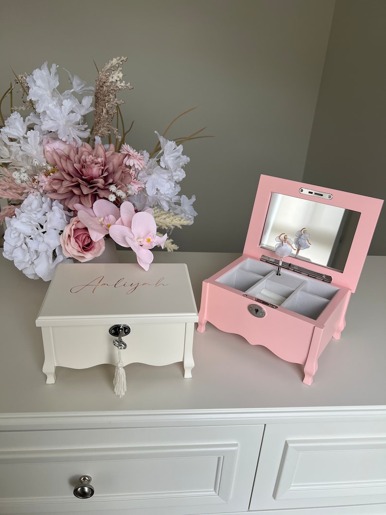 May include: Two jewellery boxes, one cream and one pink, are on a white surface. The cream box has the name "Anliyah" in cursive. The pink box has a mirror and a ballerina figurine. A bouquet of flowers is in the background.
