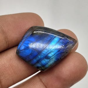 Peut inclure: Une pierre précieuse de labradorite polie avec une forme irrégulière et lisse. La pierre présente un éclat bleu irisé vibrant sur sa surface, contrasté par des zones gris foncé semi-translucides et de fines lignes de fracture naturelles.