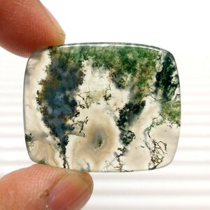 May include: A square-shaped moss agate gemstone with rounded corners. The stone features a translucent base with intricate patterns of green, white, and black inclusions, resembling a miniature landscape. The stone is held between two fingers.