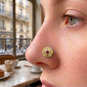 May include: A gold nose stud featuring a floral-shaped design with a central round red gemstone surrounded by four small clear stones set within the petals. The jewelry piece has an intricate, openwork metal frame and a polished gold finish.