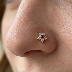 May include: A gold-colored nose stud featuring a flower design. The flower has five clear, round-cut stones surrounding a central, red gemstone. The stud is set against the skin of a nose.