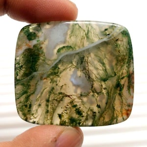 May include: A square-shaped moss agate gemstone with rounded corners. The stone features a translucent base with intricate patterns of green, brown, and white, resembling a miniature landscape. The stone is held between two fingers.