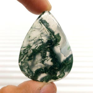 Puede incluir: Una piedra preciosa de ágata musgosa en forma de lágrima. La piedra translúcida presenta inclusiones verdes en forma de helecho dentro de una base clara. La gema se sostiene entre dos dedos, mostrando su superficie pulida y sus patrones naturales. La piedra mide aproximadamente 5 cm de largo.