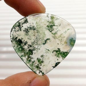 Op de afbeelding: Een hartvormige mosagaat edelsteen. De doorschijnende steen heeft witte, groene en zwarte insluitsels, die een natuurlijk, organisch patroon vormen. De steen wordt tussen twee vingers gehouden.