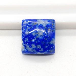 Könnte beinhalten: Ein quadratisch geschliffener, polierter Lapis-Lazuli-Edelstein. Der Stein ist ein tiefes, leuchtendes Blau mit Streifen und Flecken von hellerem Blau und Gold. Der Edelstein ist vor einem weißen Hintergrund platziert.