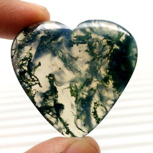 Könnte beinhalten: Ein herzförmiger Edelstein mit durchscheinendem Aussehen. Der Stein weist komplizierte Muster aus grünen und dunkelgrünen Einschlüssen auf, die Moos- oder pflanzenartigen Formationen ähneln. Der Edelstein wird von einem Finger gehalten.