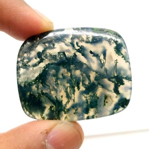 Puede incluir: Una piedra preciosa de ágata musgosa pulida, de forma cuadrada. La piedra presenta una base translúcida con intrincados patrones de verde, blanco y azul, que se asemejan a un paisaje en miniatura. La piedra preciosa se sostiene entre los dedos.