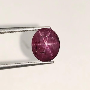 May include: A polished, oval-shaped ruby gemstone, held by metal tweezers. The ruby is a deep, rich red color with a six-ray star effect visible on its surface. The gemstone is set against a white background.